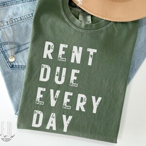 May include: An olive green t-shirt with the words "RENT DUE EVERY DAY" in white block letters. The shirt is folded and rests on a pair of light blue jeans. A tan hat and a gold necklace are also in the image.