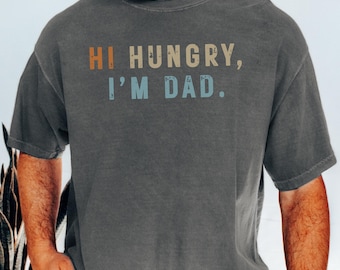 Könnte beinhalten: Dunkelgraues T-Shirt mit dem Text "Hi Hungry, I'm Dad" in braunen, beigen und blauen Buchstaben. Das Shirt ist im Comfort Colors 1717 Pepper-Stil gehalten.