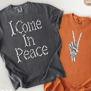 Può includere: Una t-shirt grigio antracite con il testo I Come In Peace scritto in un font bianco a forma di ossa, accanto a una t-shirt arancione che mostra una grafica bianca di una mano scheletrica che fa il segno di pace, entrambe con marchio Comfort Colors 1717.