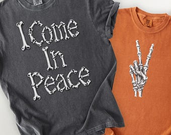 以下が含まれることがあります： 白い骨の形をしたフォントで「I Come In Peace」と書かれたチャコールグレーのTシャツと、ピースサインをする骸骨の手の白いグラフィックが描かれたオレンジ色のTシャツ。どちらもComfort Colors 1717のブランドタグが付いています。