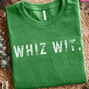 May include: A green t-shirt with the words "WHIZ WIT" printed in white. The shirt is made of a soft material and is displayed on a woven surface. The shirt also has the text "BELLA+CANVAS 3001 LEAF" printed on it.
