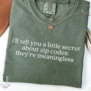 May include: An olive green t-shirt with the text "I'll tell you a little secret about zip codes: they're meaningless" in white. A gold necklace with a pendant is draped on the shirt. The shirt is labeled "Comfort Colors 1717 Moss".