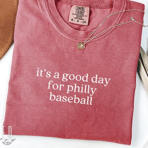 Op de afbeelding: Comfort Colors T-shirt in karmozijnrood met de witte tekst "it's a good day for philly baseball". Een gouden hangerketting ligt op de shirt. Het shirt is maat S.