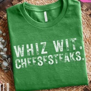 May include: Green t-shirt with the text "WHIZ WIT. CHEESESTEAKS." in distressed white lettering. The shirt is made of a soft material and has a crew neck. The brand name "BRED" is visible in the bottom left corner.