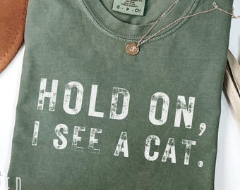 Puede incluir: Camiseta verde oliva con el texto "HOLD ON, I SEE A CAT." Un collar dorado con un colgante descansa sobre la camiseta. La etiqueta dice "COMFORT COLORS 1717 MOSS."