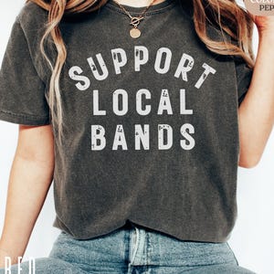 May include: Dark gray t-shirt with the text "SUPPORT LOCAL BANDS" in white block letters. The shirt is a short-sleeved crew neck style. The shirt is worn with blue jeans.