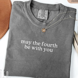 Puede incluir: Una camiseta gris con el texto "may the fourth be with you" en letras blancas. La camiseta está hecha de un material suave y cómodo y tiene un aspecto desgastado.