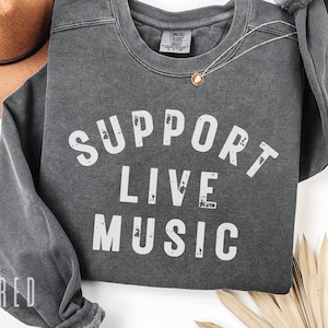 May include: A dark grey sweatshirt with the words "SUPPORT LIVE MUSIC" in distressed white lettering. The sweatshirt has a crew neck and long sleeves. A gold necklace with a pendant is draped on the shirt. A brown hat is visible in the background.