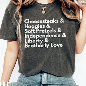 Op de afbeelding: Een donkergrijs t-shirt met witte tekst met de tekst "Cheesesteaks & Hoagies & Soft Pretzels & Independence & Liberty & Brotherly Love." Het shirt is een Comfort Colors 1717 Pepper. Het model draagt een blauwe jeans.