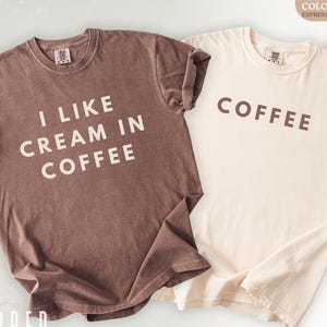 May include: Two t-shirts, one brown and one cream-coloured, with text. The brown shirt says "I LIKE CREAM IN COFFEE" in white letters. The cream shirt says "COFFEE" in brown letters. The shirts are made of a soft material.