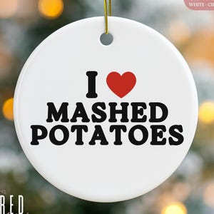 May include: A white ceramic Christmas ornament with the text "I ♥ MASHED POTATOES" in black, with a red heart symbol. The ornament has a gold string for hanging. The words "CERAMIC ORNAMENT WHITE CIRCLE" are in the upper right corner.