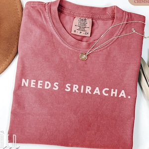 May include: Crimson red t-shirt with the text "NEEDS SRIRACHA." A silver necklace with a round pendant is draped across the shirt. The shirt has a crew neck and a tag that says "COMFORT COLORS".