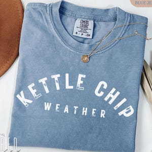 May include: A blue Comfort Colors t-shirt with the words "Kettle Chip Weather" in white block letters. A gold necklace with a pendant is draped across the shirt. The shirt is folded on a white surface.