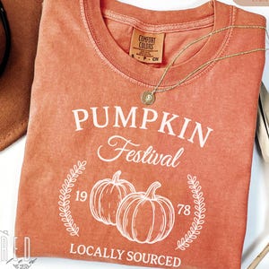 以下が含まれることがあります： 「PUMPKIN Festival」の文字と2つのカボチャのイラストが描かれた、コーラル色のTシャツ。Tシャツには「LOCALLY SOURCED」の文字と数字の「19」と「78」も表示されています。ペンダント付きのゴールドのネックレスがTシャツの上にかけられています。