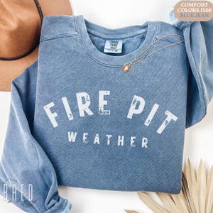 Fire Pit Weather crewneck sweatshirt in Blue Jean Comfort Colors folded flat with a hat and necklace, showcasing cozy bonfire season style.
