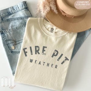 Fire Pit Weather shirt in Ivory Comfort Colors folded next to denim and a fall hat, giving neutral autumn camping outfit inspiration.