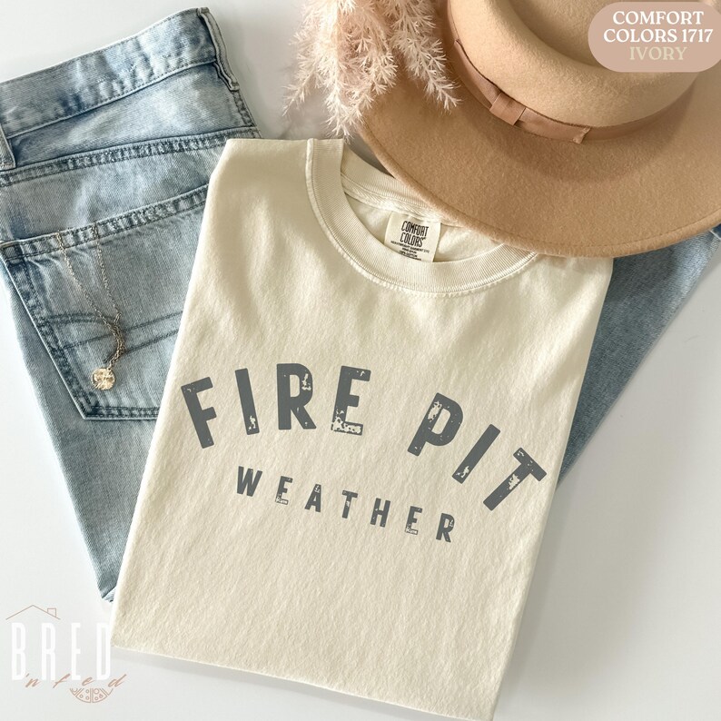 Fire Pit Weather shirt in Ivory Comfort Colors folded next to denim and a fall hat, giving neutral autumn camping outfit inspiration.