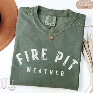 Fire Pit Weather shirt in Moss Comfort Colors tee displayed flat with a warm hat and necklace, perfect cozy fall vibe for autumn nights.