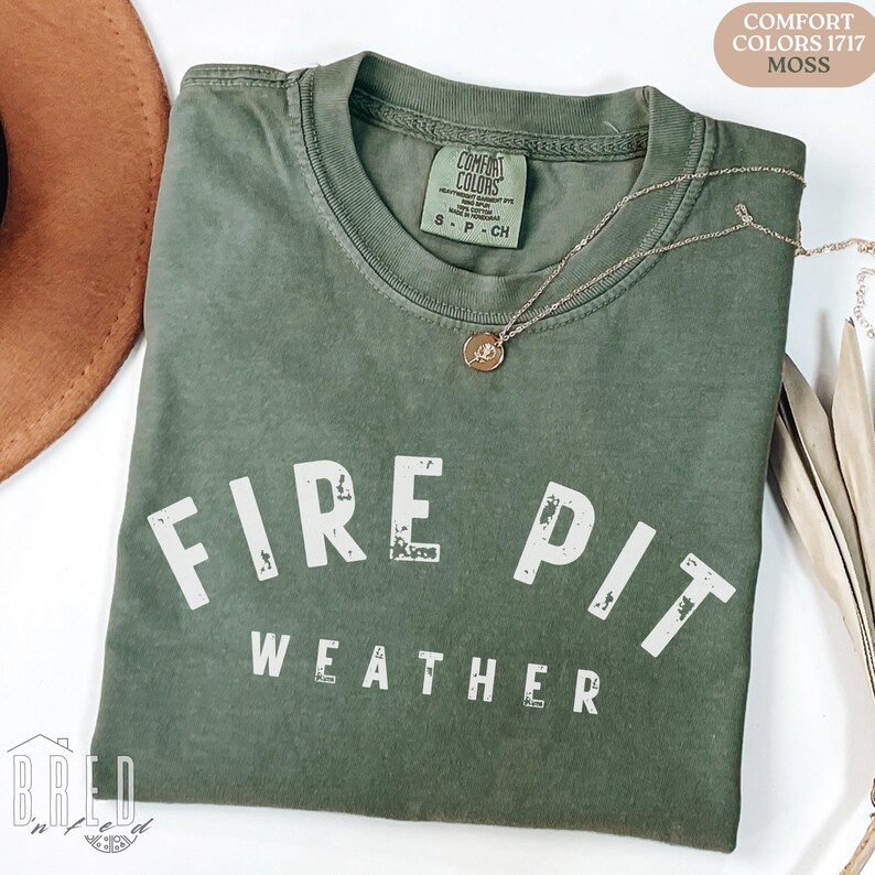 Fire Pit Weather shirt in Moss Comfort Colors tee displayed flat with a warm hat and necklace, perfect cozy fall vibe for autumn nights.