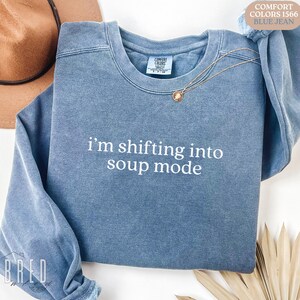 Blue Jean Comfort Colors sweatshirt that says, “i'm shifting into soup mode,” styled with gold necklace and neutral accessories. Funny Seinfeld shirt for soup fans and 90s sitcom lovers.
