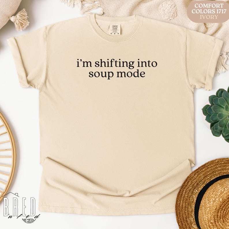 Ivory Comfort Colors shirt that says, “i'm shifting into soup mode,” flatlay with basket and greenery. Funny Seinfeld shirt, perfect gift for sitcom lovers and Soup Nazi fans.