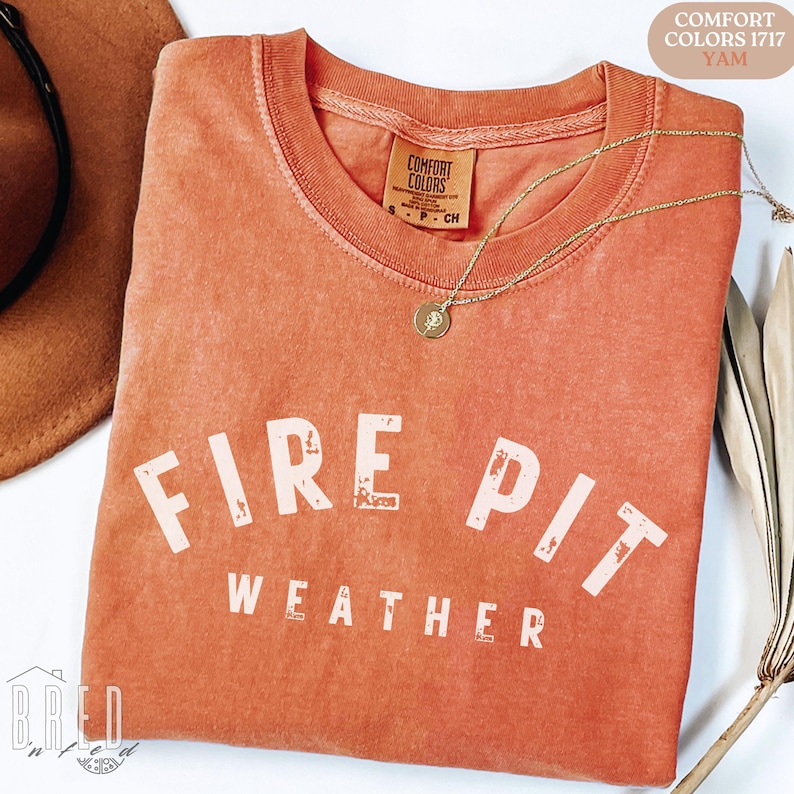 Fire Pit Weather shirt in Yam Comfort Colors folded with necklace on a warm wood background, glowing orange tones for fall bonfire season.