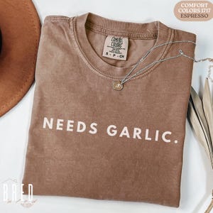 Puede incluir: Una camiseta marrón con el texto "NEEDS GARLIC." impreso en blanco. La camiseta tiene un aspecto desgastado y está hecha de una tela suave y cómoda. La camiseta es de talla S-P-CH.