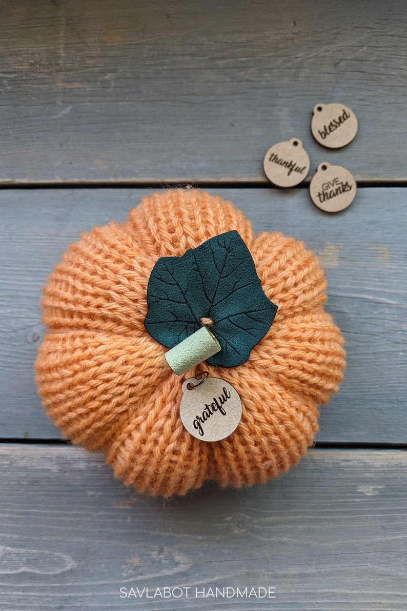 May include: A knitted orange pumpkin with a green felt leaf and a brown wooden tag that says "grateful". There are three other brown wooden tags with the words "blessed", "thankful", and "give thanks".
