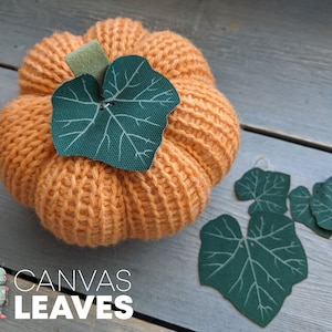 May include: A crocheted orange pumpkin with a green leaf on top. The pumpkin is surrounded by four green leaves on a wooden surface. The text "CANVAS LEAVES" is printed below the pumpkin.