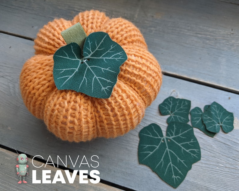 May include: A crocheted orange pumpkin with a green leaf on top. The pumpkin is surrounded by four green leaves on a wooden surface. The text "CANVAS LEAVES" is printed below the pumpkin.