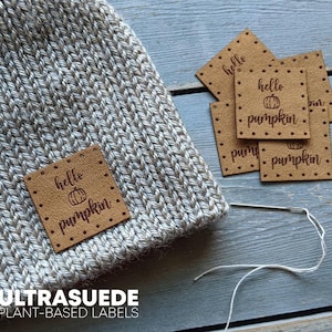 May include: A light grey knitted beanie featuring a square tan label with the text hello pumpkin and a small pumpkin illustration. Several identical tan labels are nearby, along with a silver sewing needle and white thread.