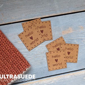 May include: A collection of square tan labels featuring the text hello world and a small heart icon. Each label has pre-punched holes along the edges for sewing. The labels are arranged near a textured, rust-colored knitted fabric item.