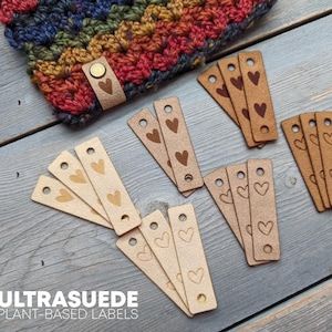 May include: A collection of Ultrasuede plant-based labels with heart designs. The labels are in various shades of brown and tan, with a button closure on a matching label attached to a colorful crocheted item. The text "ULTRASUEDE PLANT-BASED LABELS" is visible.