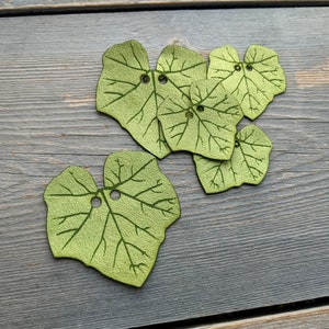 May include: Six green felt leaves with a vein pattern and a hole in the center of each leaf. The leaves are arranged in a cluster on a gray wooden surface.
