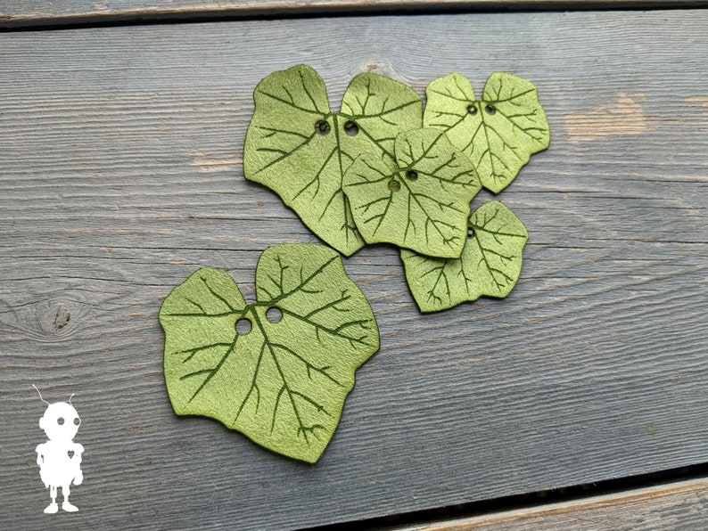 May include: Six green felt leaves with a vein pattern and a hole in the center of each leaf. The leaves are arranged in a cluster on a gray wooden surface.