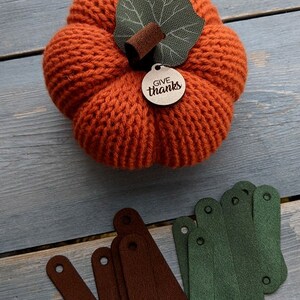 May include: A knitted orange pumpkin with a green leaf and a wooden tag that says "Give thanks". There are also several brown and green fabric strips on a wooden surface.