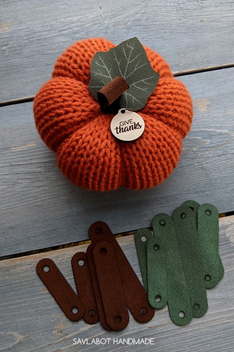 May include: A knitted orange pumpkin with a green leaf and a wooden tag that says "Give thanks". There are also several brown and green fabric strips on a wooden surface.
