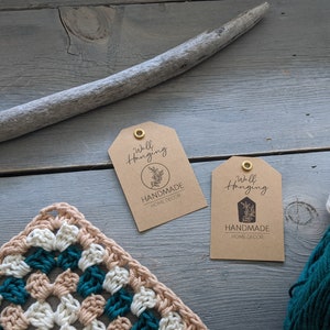Puede incluir: Dos etiquetas de papel marrón con ojales dorados, cada una con el texto "Wall Hanging" y "Handmade Home Decor" impreso en ellas. Las etiquetas están sobre una superficie de madera con un trozo de madera a la deriva y una pared de ganchillo.