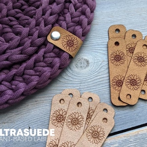 May include: A deep purple chunky knit fabric piece featuring a tan faux leather tag attached with a silver metal rivet. Several additional rectangular tan faux leather tags with laser-engraved sunflower patterns and pre-punched holes are arranged nearby.
