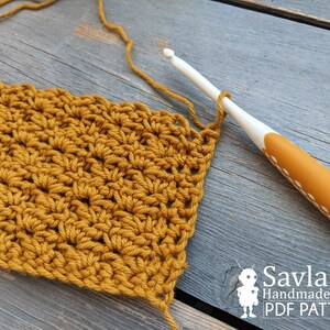 May include: A close-up of a crocheted fabric in a mustard yellow color. The fabric is being worked on with a white crochet hook. The hook has a yellow handle with white dots.