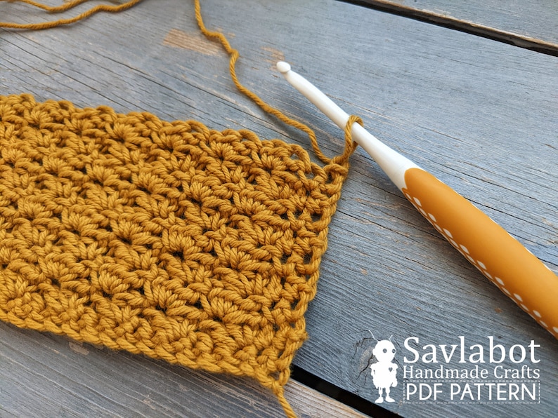 May include: A close-up of a crocheted fabric in a mustard yellow color. The fabric is being worked on with a white crochet hook. The hook has a yellow handle with white dots.