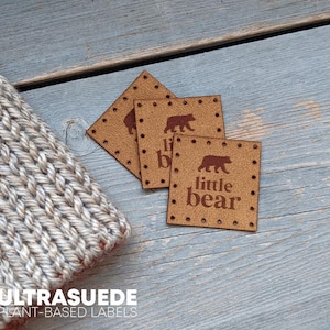 May include: Three square tan faux suede labels featuring a dark brown bear icon and the text little bear. Each label has pre-punched stitching holes along the perimeter. The labels are positioned next to a beige knitted fabric item.