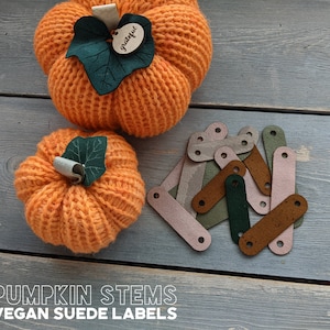 May include: A set of 10 vegan suede pumpkin stem labels in various colors, including brown, green, and pink. The labels are shaped like a stem and have a hole at the top for attaching to a pumpkin. The labels are displayed on a wooden surface with two crocheted pumpkins.