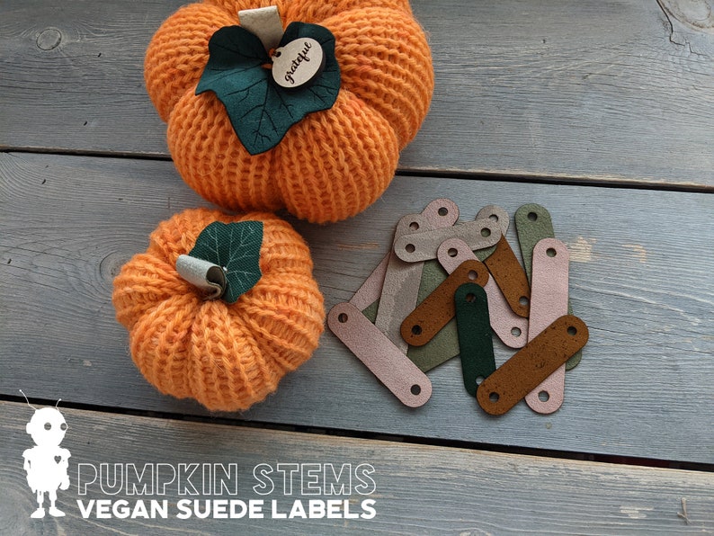 May include: A set of 10 vegan suede pumpkin stem labels in various colors, including brown, green, and pink. The labels are shaped like a stem and have a hole at the top for attaching to a pumpkin. The labels are displayed on a wooden surface with two crocheted pumpkins.