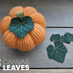 May include: A knitted orange pumpkin with a green fabric leaf on top. Four green fabric leaves are scattered on a wooden surface. The text "CANVAS LEAVES" is printed below the pumpkin.