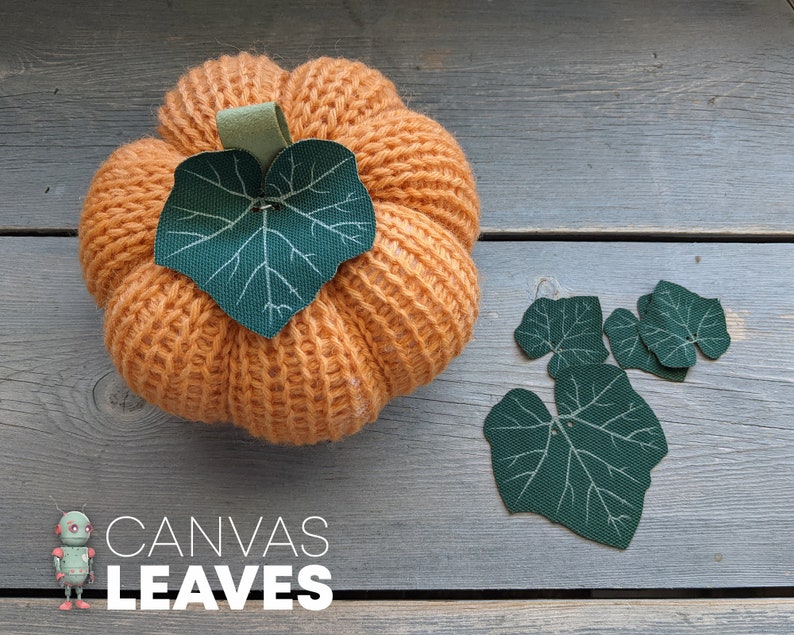 May include: A knitted orange pumpkin with a green fabric leaf on top. Four green fabric leaves are scattered on a wooden surface. The text "CANVAS LEAVES" is printed below the pumpkin.