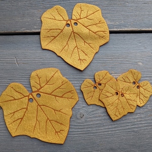 May include: Four felt leaf shapes in a mustard yellow color with brown veins. The leaves have two holes each and are arranged on a gray wooden surface.