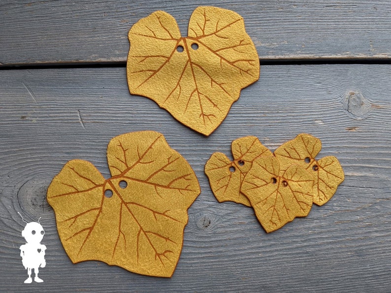 May include: Four felt leaf shapes in a mustard yellow color with brown veins. The leaves have two holes each and are arranged on a gray wooden surface.