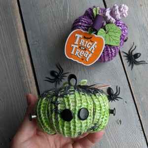 May include: Two knitted pumpkins, one green with black spiderweb details and black buttons for eyes, and one purple with a green leaf and a "Trick or Treat" tag.  The pumpkins are on a gray wooden surface with black plastic spiders.