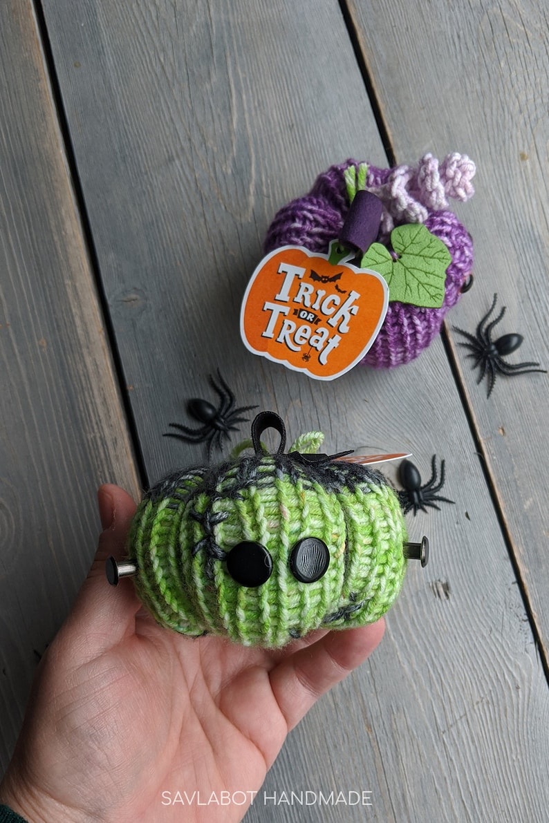 May include: Two knitted pumpkins, one green with black spiderweb details and black buttons for eyes, and one purple with a green leaf and a "Trick or Treat" tag.  The pumpkins are on a gray wooden surface with black plastic spiders.